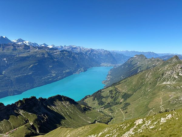 Scenic mountain landscape with railway tracks descending through green hills and a lake in the distance under a clear sky.