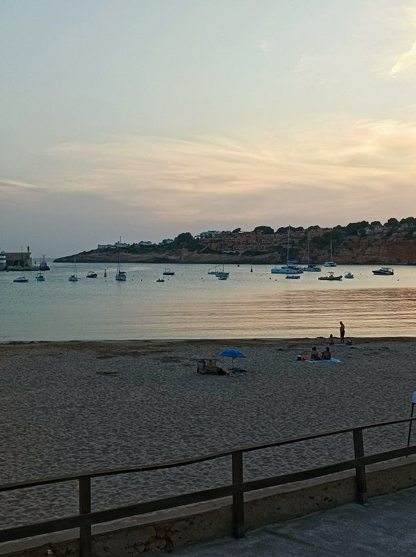 A serene beach scene at dusk with boats anchored in the water and a few people enjoying the sand.