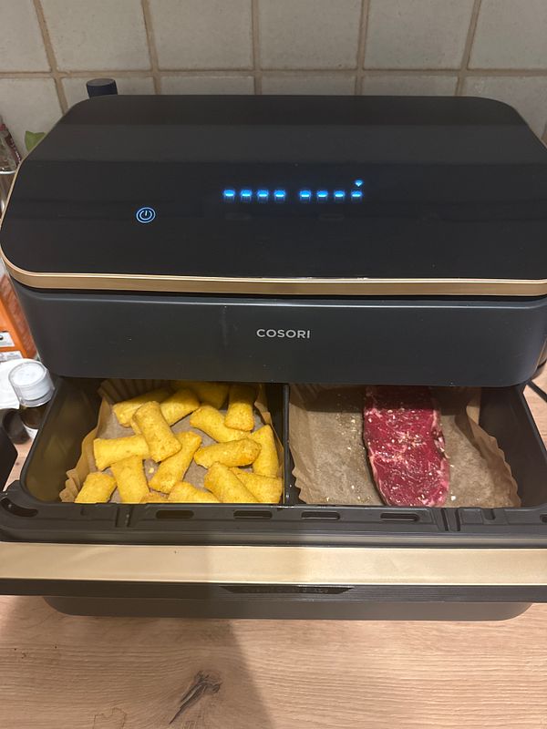 An air fryer is set up with food items inside, including frozen snacks and a piece of meat.