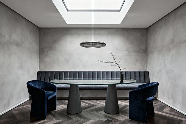 A modern dining room with a long, channel‑tufted gray banquette along the back wall and an oval glass table on two cone-shaped pedestals. Two deep blue velvet chairs sit on dark herringbone wood flooring; gray polished plaster walls, a circular smoked‑glass pendant, and a large overhead skylight provide soft light, with a small vase holding a single branch centered on the table.