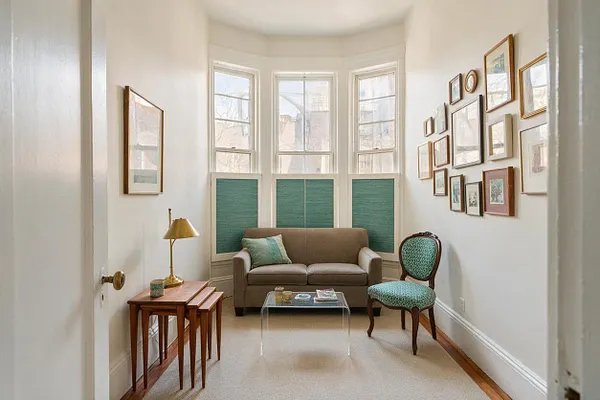 A narrow sunroom with tall bay windows, white walls, and teal window panels behind a small taupe sofa. A vintage upholstered chair, nested wooden tables with a brass lamp, a clear coffee table, and a gallery wall of framed art create an eclectic, airy seating nook.