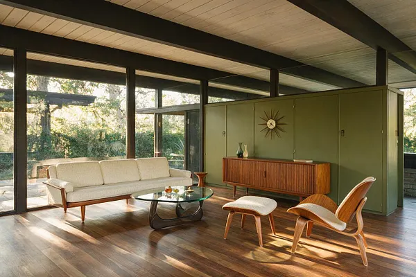 A mid-century modern front porch enclosed with floor-to-ceiling glass walls and dark exposed beams. The space features a cream upholstered sofa, curved lounge chair with matching ottoman, round glass coffee table, walnut sideboard, olive green storage wall, and warm wood flooring.