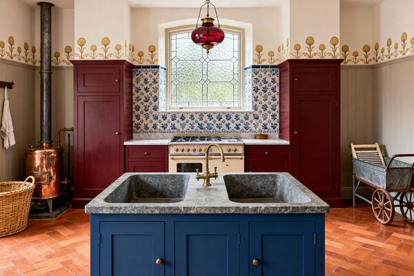 Victorian-style laundry room with a navy island topped in gray stone forming a double sink and brass bridge faucet. Terracotta herringbone tiles run under oxblood red cabinets and a cream enamel range. An arched leaded-glass window sits above blue-and-white botanical tiles. Pale paneling, a gold floral frieze, a copper boiler with iron flue, a wicker basket, and a vintage metal laundry cart add utility and period character.