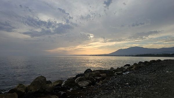 A rocky coastline with calm sea under a dramatic cloudy sky at sunset, with mountains in the distance.