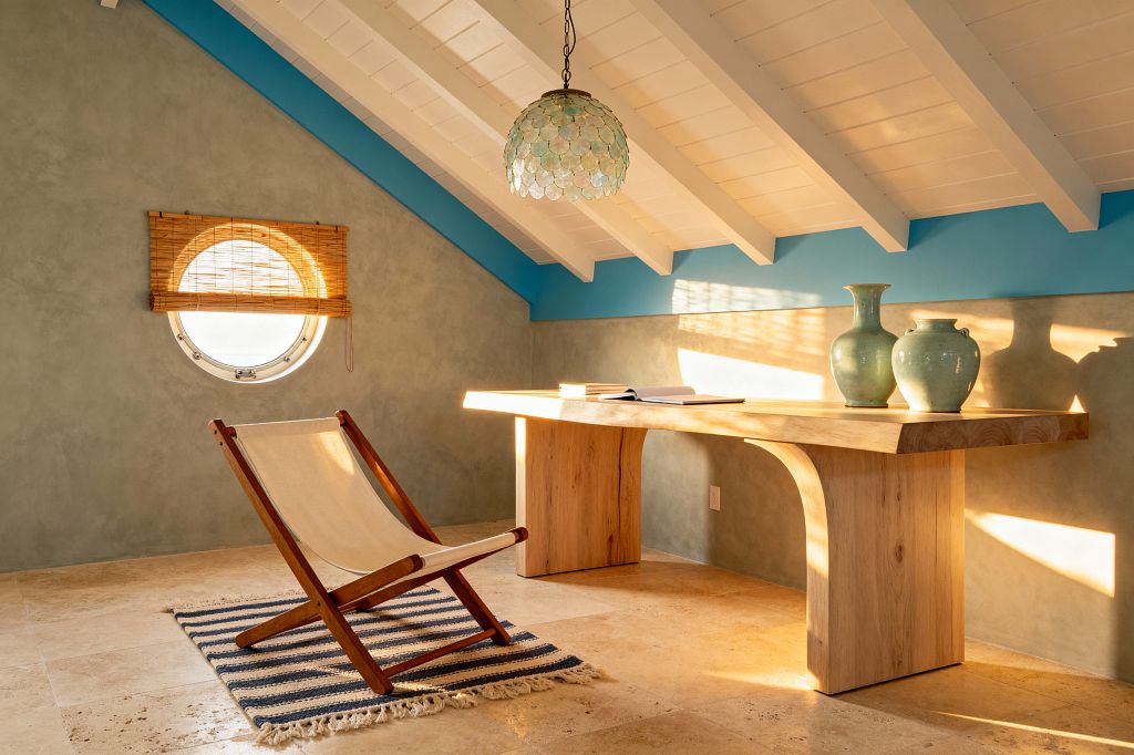 A coastal-style loft home office features a sculptural light wood desk under a white raftered ceiling with a sea-blue beam. A capiz shell dome pendant hangs overhead. On the desk sit two celadon vases and an open notebook. A canvas sling chair rests on a navy-and-cream striped rug beside a round porthole window with a bamboo shade. Sunlight washes across pale sage limewashed walls and honed travertine tile flooring.