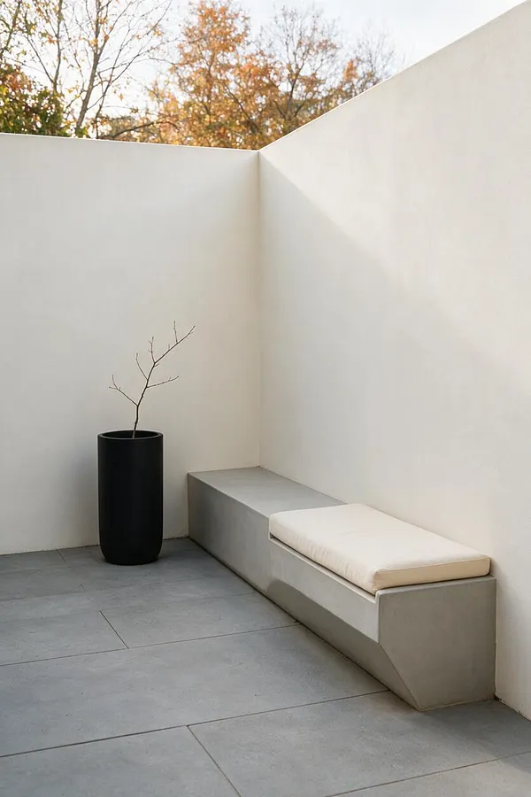 A minimalist outdoor courtyard with smooth white stucco walls, large gray stone pavers, a built-in concrete bench with a cream cushion, and a tall black planter holding a bare branch. Soft natural light and autumn trees above the wall create a calm, sparse atmosphere.