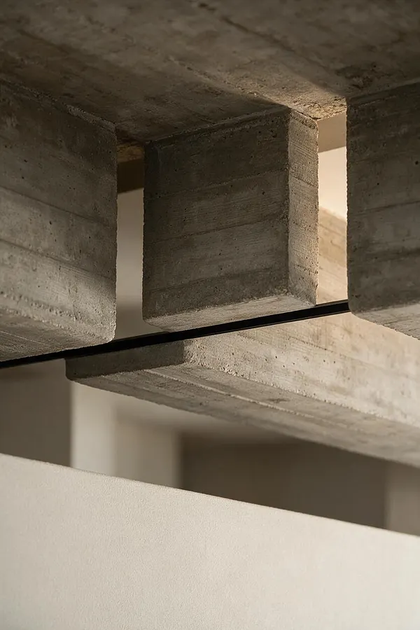 A close-up view of a Brutalist interior detail showing rough cast concrete blocks and a horizontal concrete beam with a slim dark metal line between them. Soft warm light highlights the texture of the unfinished surfaces, with a smooth pale wall visible below.