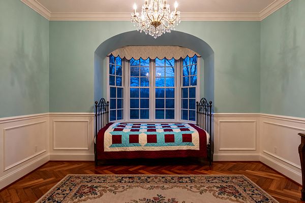 Traditional kids' bedroom with a wrought-iron daybed set into an arched bay window, topped by a scalloped valance. Walls are painted soft blue-green above white wainscoting with picture-frame molding and crown; a crystal-and-brass chandelier hangs overhead. A red, aqua, and white patchwork quilt brightens the bed, while warm herringbone hardwood floors and a floral bordered rug complete the classic look.