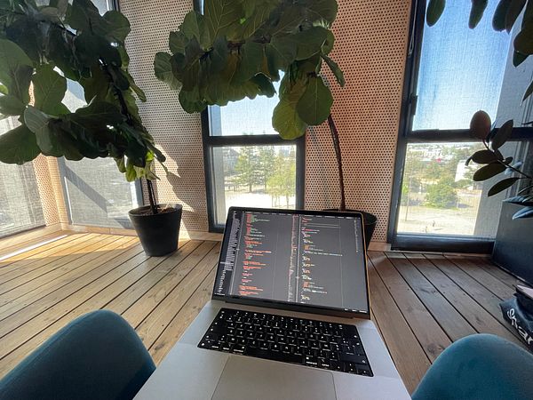 A laptop displaying code is positioned on a table in a bright, plant-filled workspace.