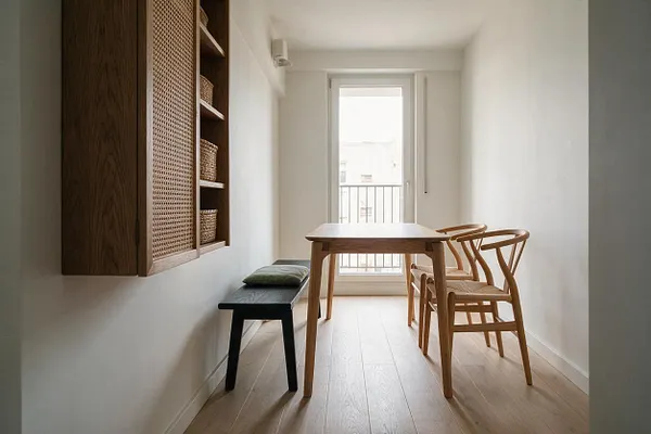 A narrow Japandi dining room with a light wood table and matching curved wooden chairs placed near a bright glass door. A dark bench with a cushion sits along the left wall beneath a wall-mounted wood cabinet with cane-front doors and woven baskets.