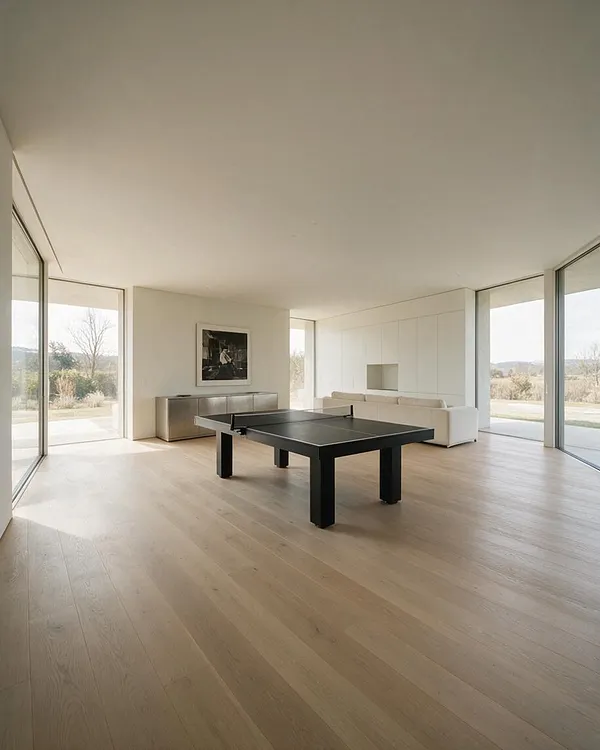 A spacious minimalist game room with a black ping pong table centered on light wood floors, a low white sofa along the back wall, and a sleek metal console beneath framed wall art. Large floor-to-ceiling glass windows on three sides fill the room with natural light and reveal a calm outdoor landscape.