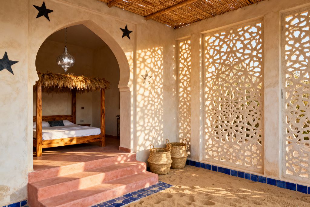 A Moroccan-inspired kids' room features an arched doorway framing a solid-wood bed topped with a palm-thatch canopy, lit by a pierced metal pendant. Carved plaster jali panels cast patterned shadows over sand-colored walls, terracotta-pink steps, and a small sandy play area edged with cobalt blue tiles. A reed ceiling and woven seagrass baskets add natural texture, while white bedding keeps the look light and airy.