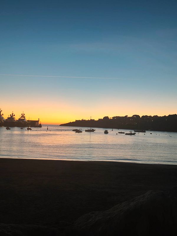 A beach at sunset with boats floating on calm water and a silhouette of a distant shoreline.