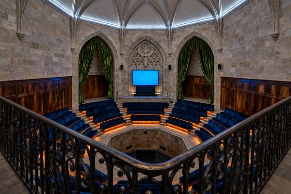 A media room set in a Gothic stone hall features ribbed vaulted ceilings, pointed arches, and carved tracery framing a flat-screen display. Tiered amphitheater seating in deep blue velvet circles the pit with amber LED step lights, emerald velvet curtains drape within side arches, dark wood wainscoting lines the walls, black wall speakers flank the screen, and a wrought-iron balcony rail frames the view.