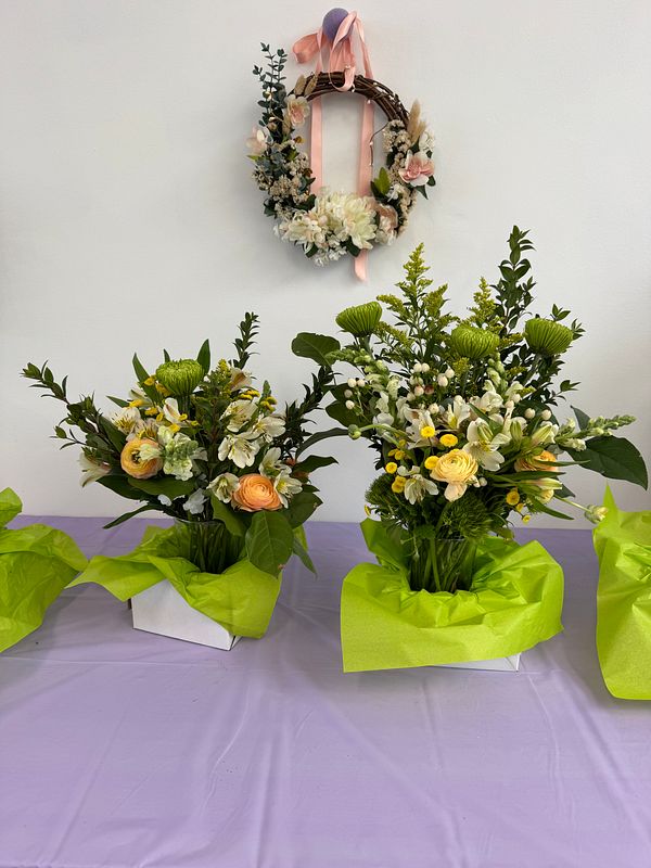 The image features two beautifully arranged flower bouquets on a table, with a decorative wreath hanging above them.