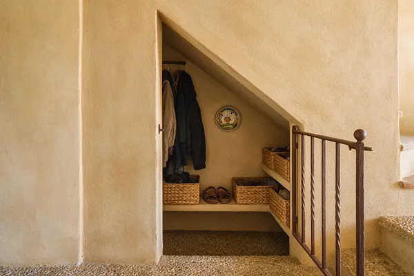 A small Mediterranean-style closet tucked under a staircase, with a sloped plaster ceiling, warm beige walls, and an open doorway framed by a simple white trim. Inside are hanging coats, woven wicker storage baskets, a pair of sandals, and a decorative ceramic plate on the back wall, with a dark metal stair railing in front.