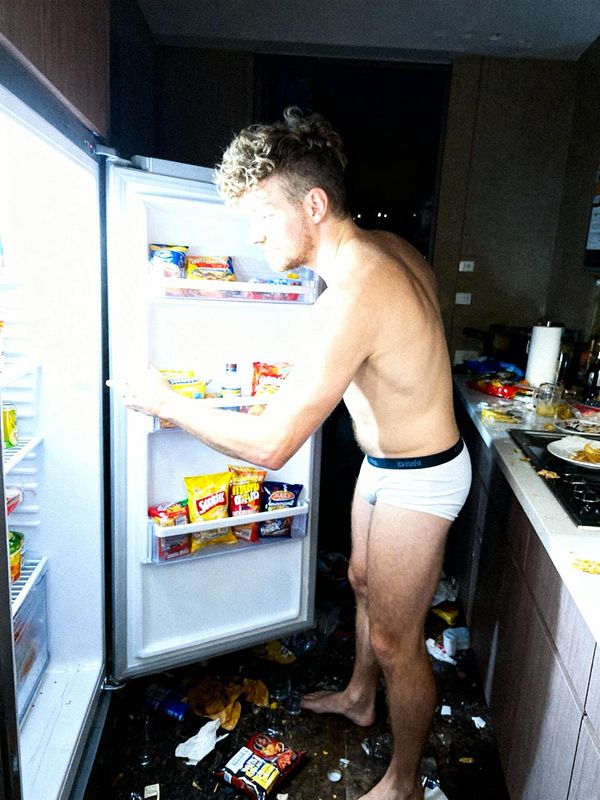 A man in his underwear is rummaging through an open refrigerator in a messy kitchen.