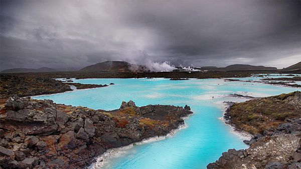 A stunning geothermal landscape featuring bright blue waters surrounded by dark volcanic rock under a moody sky.