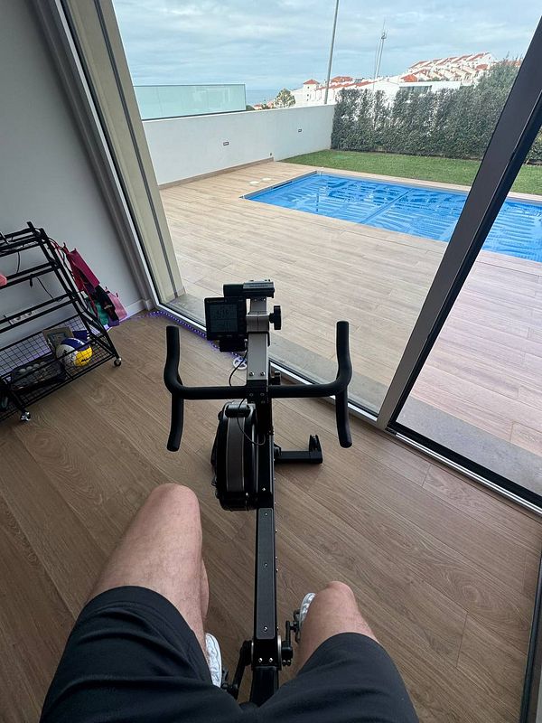 A person is seated on a rowing machine with a view of a swimming pool and outdoor area.