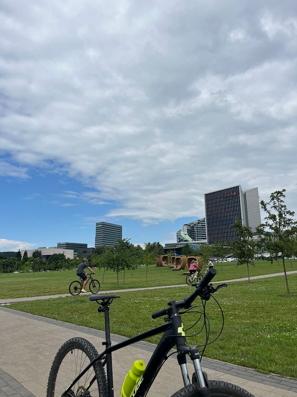 A scenic park view featuring cyclists and modern buildings under a partly cloudy sky.