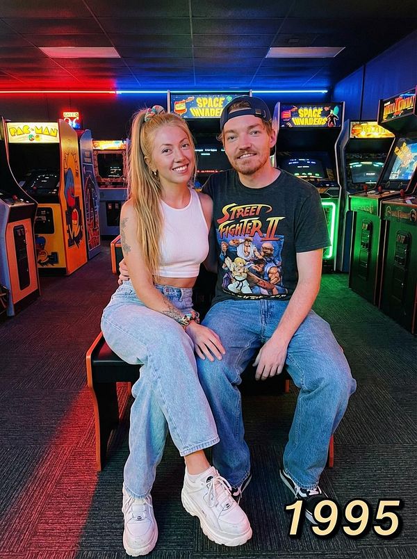 A vertical photo of a young couple lying close together on a bed, smiling and holding hands, surrounded by cozy bedding and pillows.