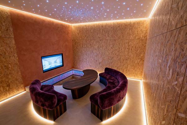 A contemporary media room with OSB wood wall panels and a terracotta plaster feature wall holds a flush‑mounted TV. Two curved sofas in deep plum velvet encircle a kidney‑shaped dark‑wood table on a smooth light microcement floor. Warm LED strips trace the ceiling, baseboards, and seat bases, while a starry fiber‑optic ceiling and a low bench with pixel‑style LEDs create an immersive, softly lit atmosphere.
