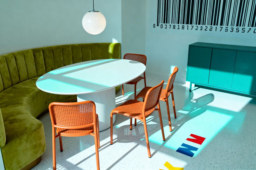 A bright dining nook features a curved olive-green velvet banquette wrapping a white oval table with a fluted pedestal base. Four tangerine perforated chairs sit opposite under a white globe pendant. A teal sideboard stands against a white wall displaying a large black barcode graphic. Sunlight washes across a light speckled terrazzo floor with inlaid primary-color letters, casting crisp shadows and emphasizing the room’s bold, graphic forms.