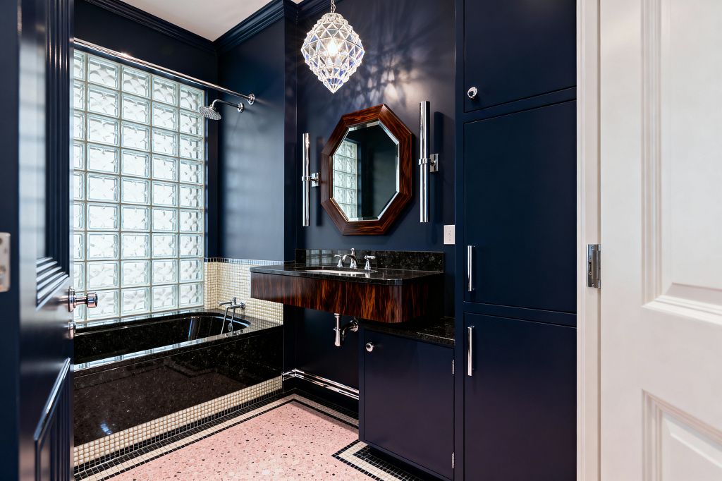 An Art Deco bathroom with deep navy walls and cabinetry, a floating wood-veneer vanity topped with black granite, and an octagonal wood-framed mirror flanked by tall chrome sconces beneath a faceted crystal pendant. A tub-shower with black stone surround and cream mosaic trim sits before a large glass-block window, while pink terrazzo flooring with a black-and-white border and polished chrome fixtures complete the space.