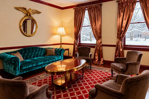 A Traditional living room features a teal tufted velvet sofa, three taupe velvet armchairs, and an oval mahogany coffee table with brass trim on a red lattice wool rug. Cream walls with burgundy chair rail and window frames hold copper satin drapes, a brass floor lamp, and a gilded round mirror topped by an eagle; snowy trees are visible through the windows, and warm lighting creates a formal conversation area.