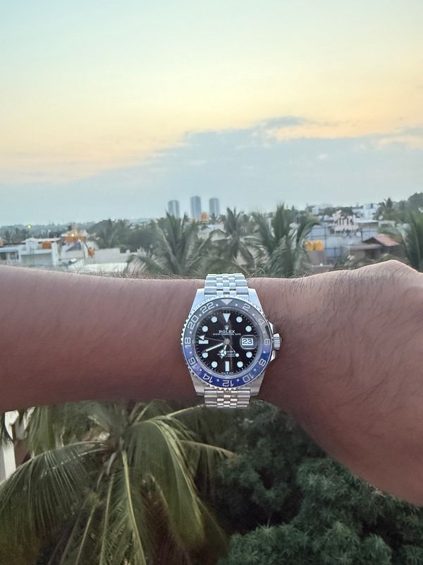 A hand wearing a Rolex watch is extended towards a sunset view over a cityscape.