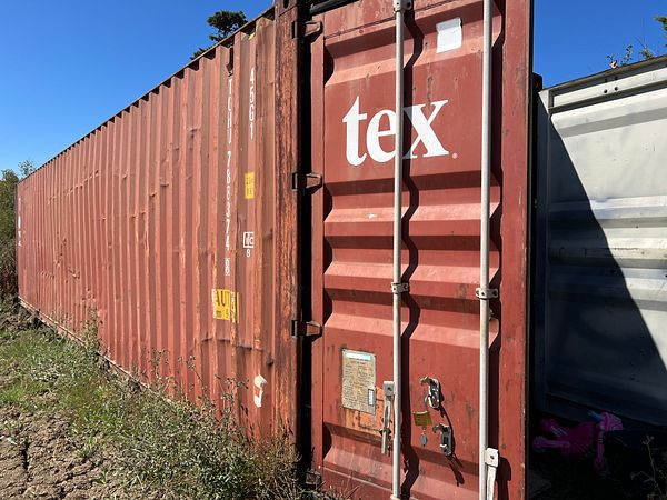 A weathered shipping container is positioned outdoors, surrounded by vegetation.