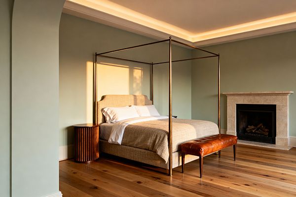 A serene bedroom with soft sage walls features a brass four-poster bed upholstered in beige linen with white bedding. A cognac leather bench sits at the foot, a fluted dark-wood round nightstand is beside the bed, and a pale limestone fireplace anchors the corner; wide-plank oak flooring and warm cove lighting along the ceiling create a gentle glow, with late-afternoon sunlight casting shadows across the room.