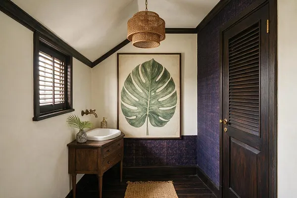 A small tropical-style powder room with a dark wood vanity, white vessel sink, and brass faucet beneath a framed oversized green leaf print. The room features a woven rattan pendant light, dark trim and louvered door, deep blue textured wall covering, and a natural jute rug on dark wood flooring.