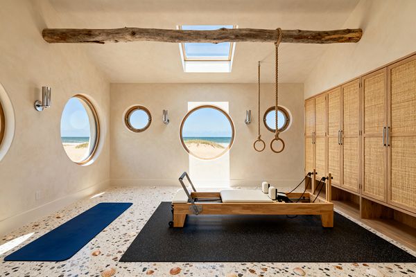 A serene coastal home gym with sandy limewashed walls shows a wood Pilates reformer on a black rubber mat and a blue yoga mat on terrazzo flooring inlaid with seashells. Round porthole windows and a larger central circular window reveal dunes and ocean; a skylight above and chrome sconces provide additional light. A driftwood ceiling beam suspends rope gymnastic rings, and a wall of oak cabinets with woven rattan doors lines the right side.