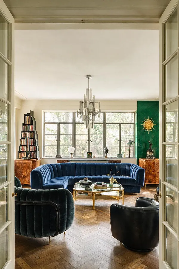 A bright sunroom styled in Art Deco fashion, featuring a deep blue curved velvet sofa, two dark lounge chairs, a brass-and-glass coffee table, and a dramatic tiered chandelier. Large paned windows wrap the room, while rich wood flooring, amber burl wood cabinets, and a green accent wall with a gold sunburst add warmth and glamour.