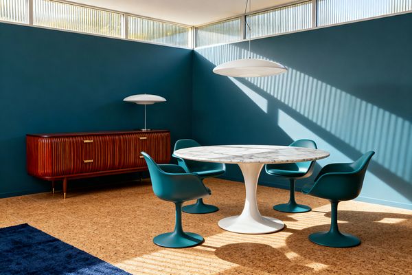 A Mid-Century Modern dining room with deep teal walls features a white saucer pendant above a round table with a white pedestal base and gray-veined marble top, surrounded by four teal tulip armchairs. To the left, a fluted reddish wood credenza with brass pulls supports a white mushroom lamp, while clerestory windows with ribbed glass cast diagonal shadows across the cork floor and a navy rug peeks into the foreground.