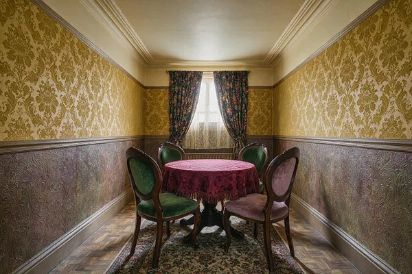 A narrow Victorian-style basement dining room with ornate gold damask wallpaper, dark embossed wainscoting, and a small round table covered in a burgundy cloth. Four upholstered wood chairs surround the table beneath floral drapes framing a back window, while a patterned rug and parquet floor complete the period look.