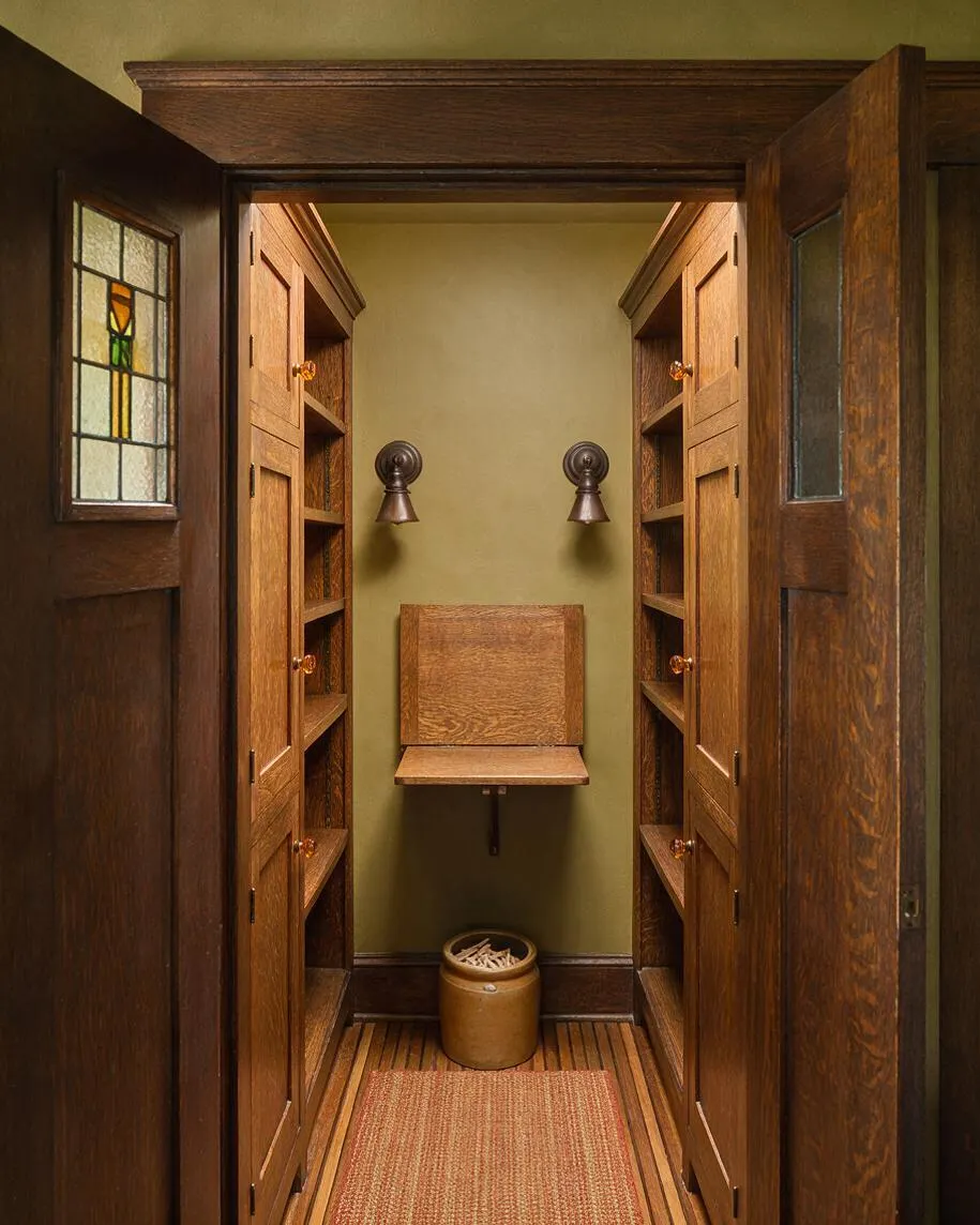 A narrow Craftsman-style closet with rich wood built-in storage on both sides, paneled doors with small stained-glass windows, and brass-toned hardware. The back wall is painted a muted olive tone and features two wall sconces, a fold-down wood bench, a small container on the floor, and a woven runner on the floorboards.