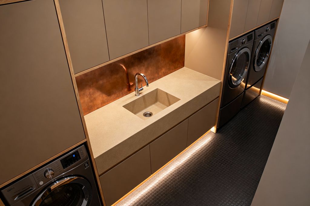 Angled view of a modern laundry room featuring flat taupe cabinetry edged in light wood, a sand-beige solid-surface countertop, and an integrated rectangular sink with a chrome faucet. A patina copper sheet forms the backsplash. Stainless front-loading washer and dryer are built into tall cabinet bays. The floor is dark charcoal penny tile. Warm LED strip lighting glows along the toe-kick and niches, creating a clean, streamlined atmosphere in the narrow space.