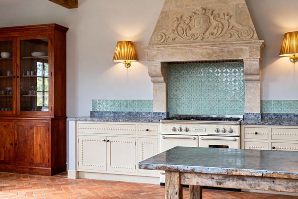 A traditional kitchen features a cream enamel range set beneath a grand carved limestone hood. Glossy seafoam tiles with a raised motif line the backsplash above blue-gray granite counters and cream cabinets detailed with bead molding and dark knobs. Brass wall sconces with pleated gold shades flank the hood. At left stands a tall glass-front hutch in warm wood. The floor is herringbone terracotta, and a rustic table with a weathered zinc top sits in front.