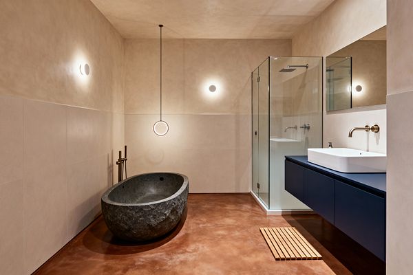 A minimalist modern bathroom features a rough-hewn oval stone soaking tub beside a freestanding brushed-brass filler, a slim ring pendant, and two small wall sconces. Opposite, a frameless glass shower and a floating matte-navy vanity with a white vessel sink sit against warm beige plaster and large-format tile walls, above a terracotta-toned polished concrete floor with a slatted wood bath mat and a wide mirror reflecting the soft, warm light.