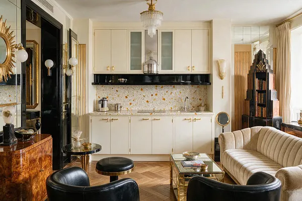 A compact Art Deco studio apartment with a cream kitchenette centered on a terrazzo backsplash, glossy black trim, and brass cabinet hardware. The seating area includes a curved beige velvet sofa, black leather chairs, mirrored and glass tables, a sunburst mirror, and decorative vintage-style lighting.