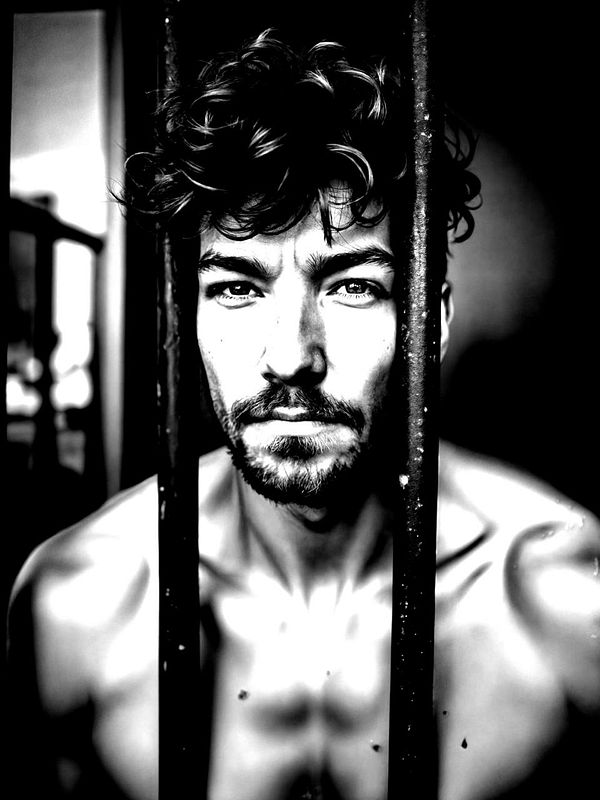 A black and white portrait of a man with curly hair, looking intensely through bars.