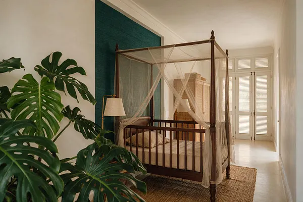 A tropical-style nursery with a dark wood canopy crib draped in sheer mosquito netting, set beside a teal accent wall and a woven rug. Large monstera plants fill the foreground, while a wicker storage cabinet and bright shuttered doors add warmth and natural texture.