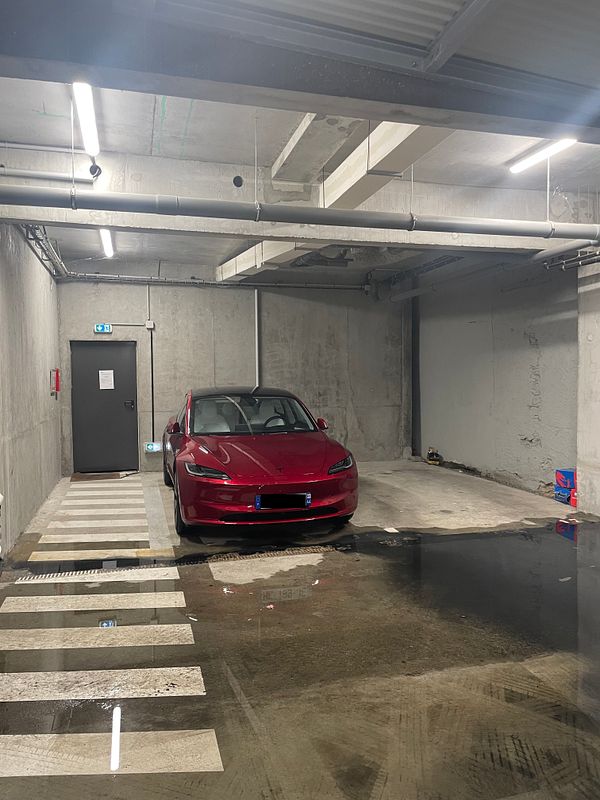 A clean white Tesla Model 3 parked in a garage with a red toolbox and a hose visible on the right side.
