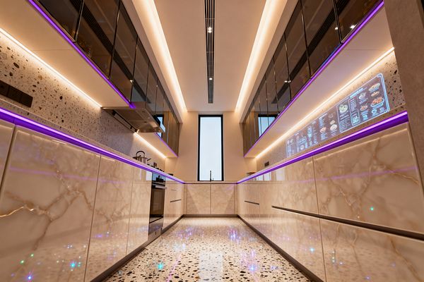 A long, narrow galley kitchen features glossy marble-effect base cabinets and smoked-glass upper units flanking a terrazzo floor and backsplash. Warm white cove lighting and under-cabinet LEDs with a purple accent strip illuminate handleless drawers, a stainless range hood, and a central sink below a tall black-framed window. A digital touchscreen panel spans part of the right backsplash, reinforcing the sleek, futuristic aesthetic.
