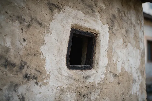 A close-up view of a weathered exterior wall in muted earth tones, finished with cracked plaster and visible stains. A small square window with a dark wooden frame sits near the center, emphasizing the raw, handmade character of the facade.