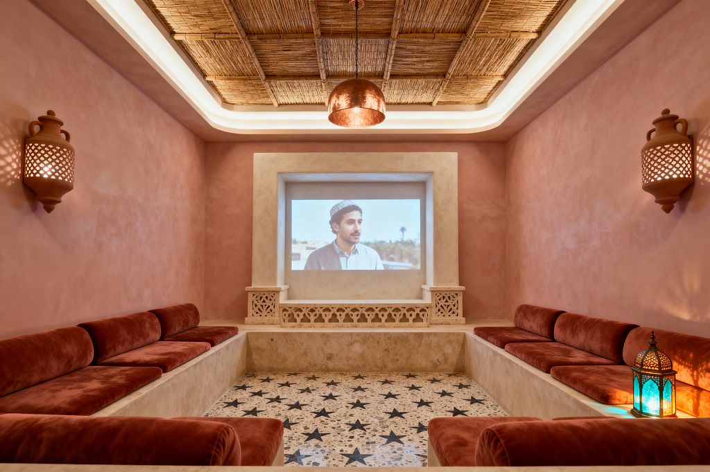 A Moroccan-style media room with blush plaster walls features a sunken U-shaped built-in bench topped with rust velvet cushions and a projector screen set into a sandy plaster niche with carved lattice details. A terrazzo floor with black star inlays, woven reed ceiling with cove lighting, terracotta pierced sconces, a copper dome pendant, and a small colored-glass lantern create a warm, artisanal atmosphere.