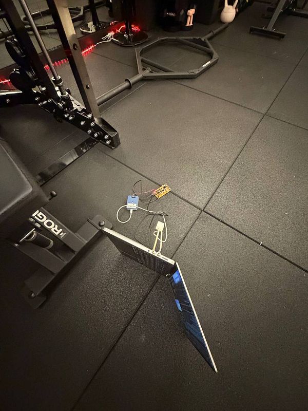 A laptop connected with wires to electronic components is placed on a gym floor near exercise equipment.