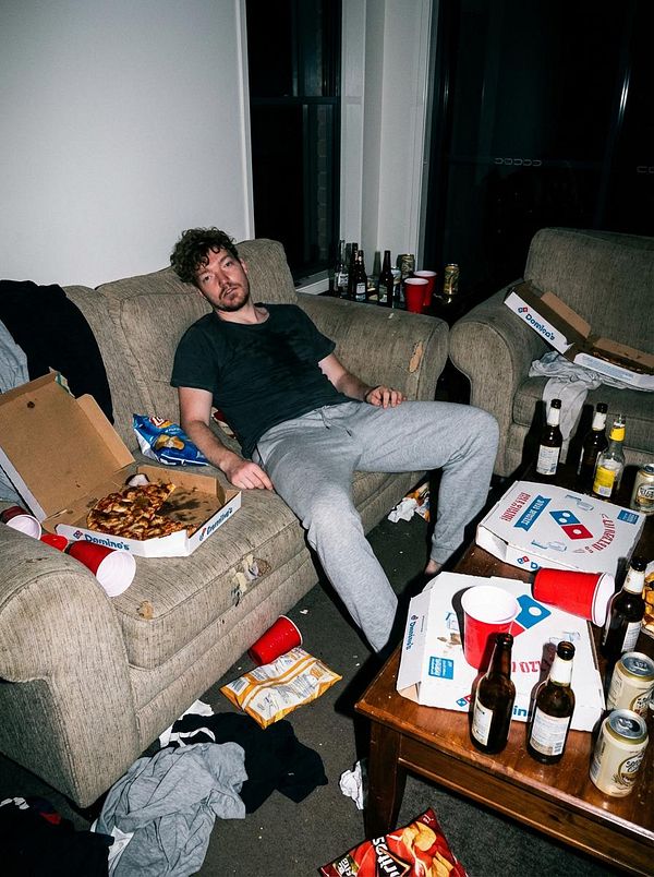 A disheveled man sits on a messy couch surrounded by empty bottles and food remnants.
